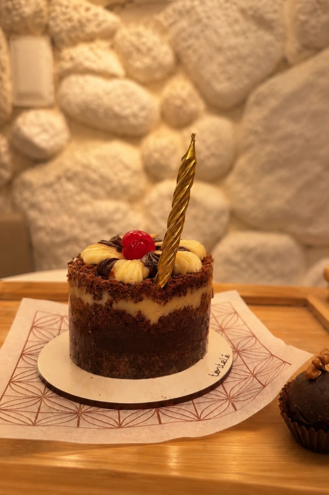 Bolinho de aniversário com vela dourada sobre uma bandeja de madeira, em frente a uma parede de pedra branca